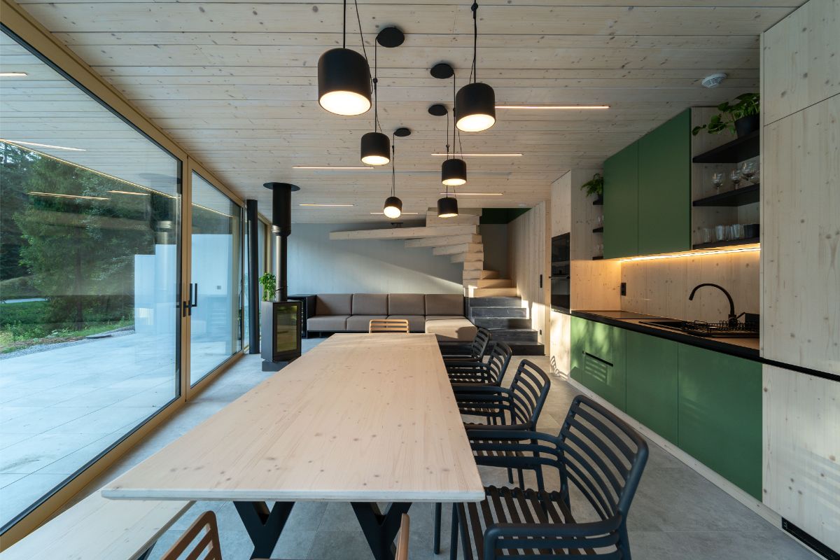 Interiér so svetlým dreveným stropom, čiernymi závesnými lampami, dreveným schodiskom a presklenou stenou s výhľadom na les.