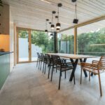 Svetlý moderný interiér s dlhým dreveným stolom, presklenými obvodovými stenami, zelenou linkou a priznaným dreveným obkladom.