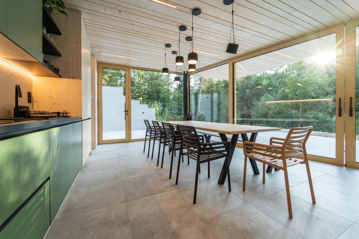 Svetlý moderný interiér s dlhým dreveným stolom, presklenými obvodovými stenami, zelenou linkou a priznaným dreveným obkladom.