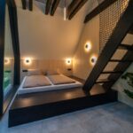 Moderná spálňa s čiernym dreveným pódiom, kruhovými nástennými svietidlami a svetlým obkladom.