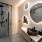 Kúpeľňa s organickými podsvietenými zrkadlami, tmavým kamenným umývadlom a svetlým dreveným panelovaním.