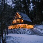 Osvetlená moderná drevostavba s ostrou čiernou šikmou strechou a preskleným štítom v zasneženom lese počas zimného večera.