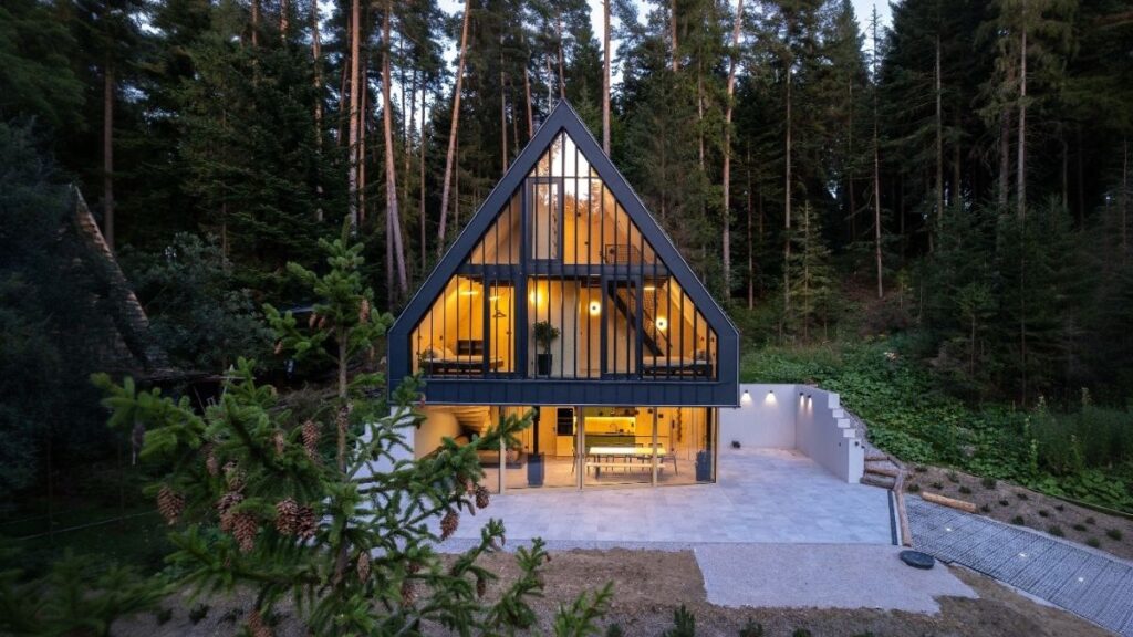 Večerný exteriér chaty s čiernou konštrukciou, preskleným priečelím, dláždenou terasou a osvetleným minimalistickým interiérom.
