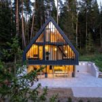 Večerný exteriér chaty s čiernou konštrukciou, preskleným priečelím, dláždenou terasou a osvetleným minimalistickým interiérom.