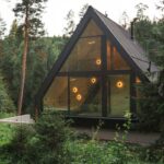 Čierna kovová konštrukcia fasády s veľkými sklenenými tabuľami, odrážajúcimi okolitý les a žlté vnútorné bodové osvetlenie.