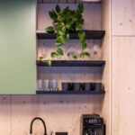 Kuchynský kút so zelenými matnými skrinkami, čiernym drezom, svetlým dreveným obkladom stien a otvorenými čiernymi policami.