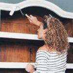 Mladá žena maľuje valčekom starožitnú skriňu nabielo.