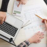 Pohľad zhora na stôl s pôdorysom domu, notebookom a kalkulačkou.