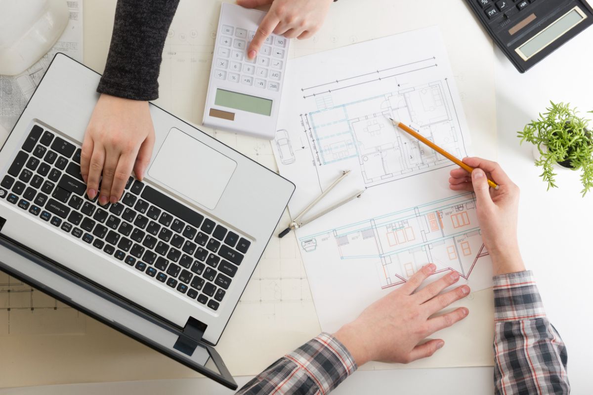 Pohľad zhora na stôl s pôdorysom domu, notebookom a kalkulačkou.