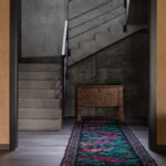 Chodba s farebným kobercom, vyrezávanou drevenou truhlou a betónovým schodiskom s čiernym zábradlím.