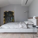 Svetlá moderná spálňa v odtieňoch sivej s drevenou podlahou a posteľou s károvaným poťahom pri abstraktnom obraze.