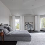 Svetlá podkrovná spálňa so šedou manželskou posteľou, vysokým kobercom a veľkými bielymi oknami.