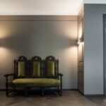 Baroková tmavohnedá lavica so zeleným zamatovým čalúnením pred sivou stenou s lištami a dvomi nástennými lampami.