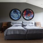 Svetlá spálňa s drevenou podlahou, dvoma sivými lôžkami a dvoma kruhovými abstraktnými obrazmi na stene.