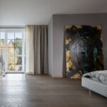 Moderná spálňa s drevenou podlahou, sivými stenami a veľkým abstraktným obrazom pri sklenenej stene s dverami.