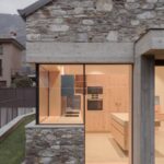 Kamenný dom s presklenou stenou, betónovými prvkami a kuchyňou s drevenými skrinkami.