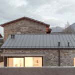 Vonkajší pohľad na kamenný dom s plechovou strechou a veľkými oknami s výhľadom na hory.