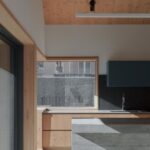 Kuchyňa s drevenými skrinkami, betónovým ostrovčekom a tmavomodrou závesnou skrinkou pod dreveným stropom.