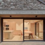 Pohľad cez presklenú stenu na interiér kuchyne a jedálne s dreveným nábytkom a červenými stoličkami.