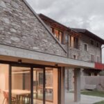 Kamenný dom s modernou betónovou prístavbou a veľkými presklenými dverami s výhľadom na trávnik.