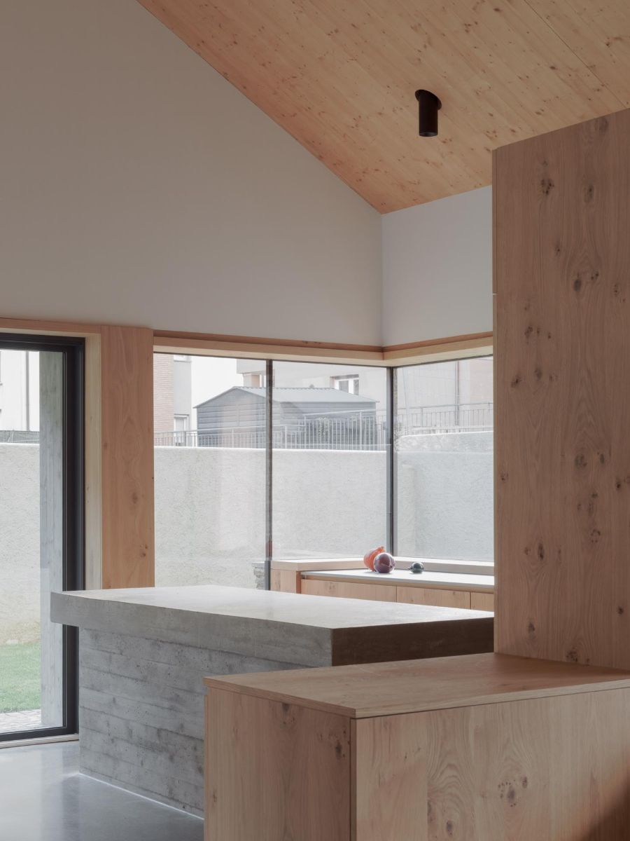 Interiér s dreveným šikmým stropom a betónovou kuchynskou linkou s drevenými skrinkami pri veľkom okne.