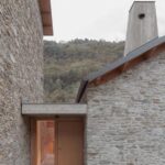 Vchod do kamenného domu s drevenými dverami a drevenou strechou vedľa vysokého betónového komína.