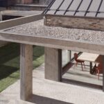 Pohľad zhora na terasu s drobným štrkom, betónovým trámom, kamennou dlažbou a presklenou jedálňou.