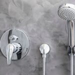 Detail na sprchu a batériu na stene so sivou stierkou.