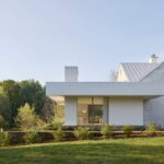 Biela minimalistická prístavba s plochou strechou a presklením vedľa domu s kamennou fasádu a sedlovou strechou.