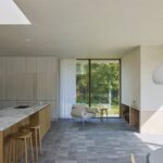 Kuchynský ostrov s mramorovanou doskou, drevené barové stoličky a svetlé kreslo v otvorenom interiéri.