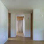 Moderná minimalistická chodba s bielymi stenami a drevenou podlahou, vedúca k priestoru so závesom.