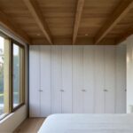 Spálňa s dreveným trámovým stropom, vstavanými bielymi skriňami a veľkým oknom.