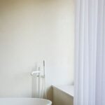 Voľne stojaca biela vaňa s modernou vysokou batériou s jemným bielym závesom a štruktúrovanou omietkou.