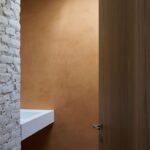 Pohľad cez otvorené dvere do kúpeľne s bielym umývadlom a terakotovou stierkou.