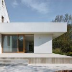 Moderná biela prístavba s betónovou terasou, posuvnými dverami a pôvodnou kamennou fasádou domu.