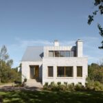 Biely kamenný dom so sivou plechovou strechou, drevenými oknami a betónovými schodmi v záhrade.