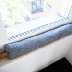 Sivý textilný vankúš proti prievanu na mramorovom parapete pod bielym plastovým oknom.