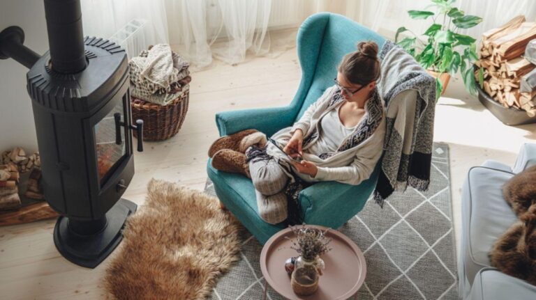 Žena v pletenom svetri a hnedých papučiach sediaca v tyrkysovom kresle pri čiernej liatinovej peci.