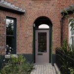 Dom s tehlovou fasádou s klenutým vchodom, tlmené hnedé dvere s presklením a svetlá kamenná dlažba v predzáhradke.
