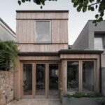 Úzky dom s dreveným obkladom, presklenou stenou s dverami a betónovým kvetináčom s rastlinami.