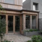 Drevená prístavba s presklenými dverami a betónovým záhonom pri tehlovej terase.