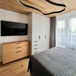 Svetlá spálňa s dreveným stropom, bielym šatníkom, dubovou komodou a moderným čiernym LED svietidlom.