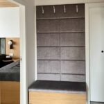 Predsieň s hnedým čalúneným panelom, striebornými vešiakmi a dubovou lavicou na čiernych kovových nohách.