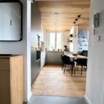 Pohľad z chodby na svetlú kuchyňu s dreveným stropom, sivou linkou a moderným jedálenským kútom.