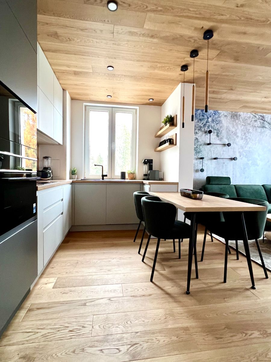 Kuchynský kút so sivým nábytkom, drevenou pracovnou doskou, bielym oknom a visiacimi čiernymi svietidlami.