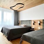 Manželská posteľ s tmavosivou pokrývkou, zeleným čalúneným čelom a dubovým stenovým obkladom s bielymi lampami.