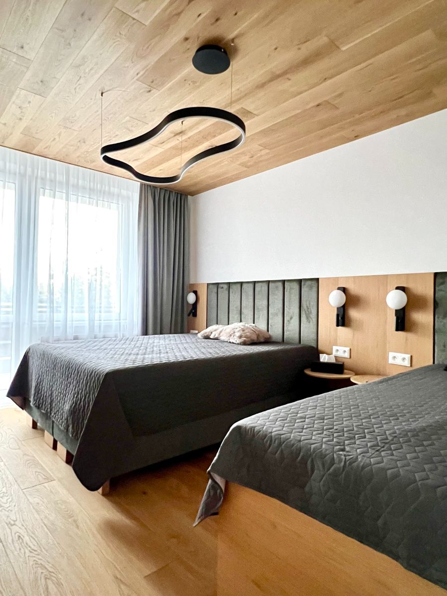 Manželská posteľ s tmavosivou pokrývkou, zeleným čalúneným čelom a dubovým stenovým obkladom s bielymi lampami.