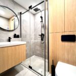Moderná kúpeľňa so skleneným sprchovacím kútom, čiernou batériou a dreveným dekorom na skrinkách aj stene.