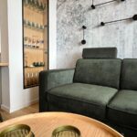 Zelená látková sedačka pri bielej stene s presklenou vitrínou a minimalistickým čiernym nástenným osvetlením.