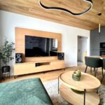 Obývacia stena z prírodného dreva s veľkým televízorom, čiernymi reproduktormi a okrúhlym konferenčným stolíkom.